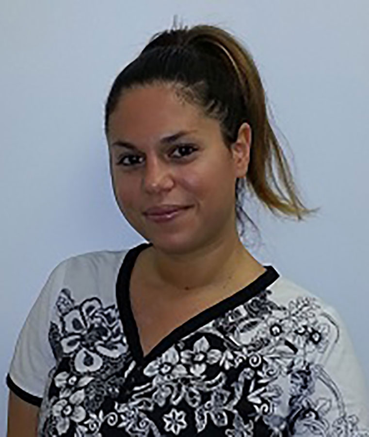 A woman with a ponytail, wearing a black shirt and white pants, poses against a plain background.