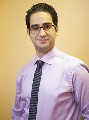 A man in a purple shirt, wearing glasses and a tie, stands against a light wall.