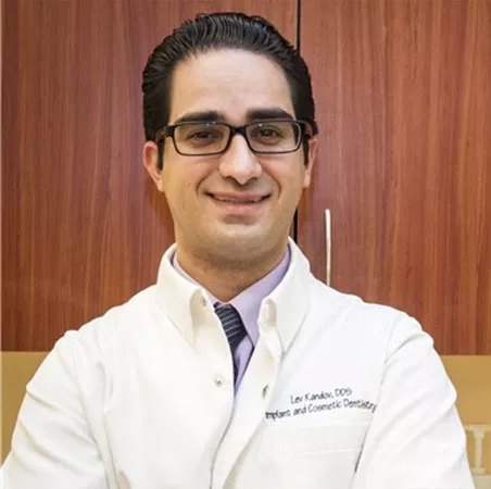 The image shows a man standing with his arms crossed, wearing glasses, a white shirt, and a name tag, posing in front of a wooden cabinet with a glass door that has text on it. He is smiling and appears to be in a professional setting, possibly an office or a dental clinic.