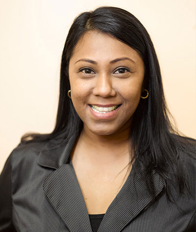 The image shows a woman with a smile, wearing a dark top and black pants, against a light background.