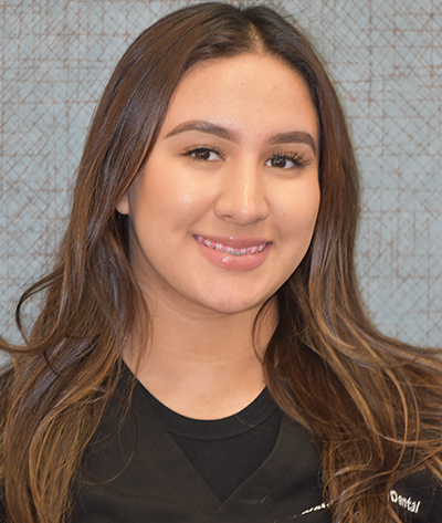 The image shows a person smiling at the camera with their hair styled down. They are wearing makeup and have a visible earring. The individual appears to be indoors, possibly in an office setting, indicated by the presence of a patterned wall behind them.