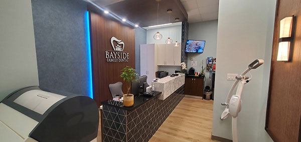 The image shows an interior space that appears to be a modern dental clinic or office with a clean and contemporary design, featuring a reception area with seating, a counter with a sink, a mirror, and a desk with a computer monitor.