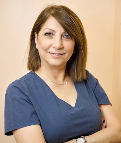 The image shows a woman wearing a blue shirt and standing with her arms crossed. She has short hair and is smiling slightly towards the camera.