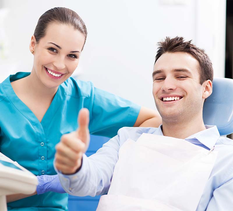 The image is a photograph of a dental clinic setting, featuring a male patient giving a thumbs-up sign and a female dental professional standing beside him.