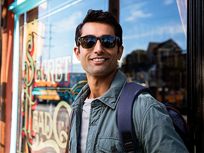 The image features a man standing outdoors, wearing sunglasses and a backpack, posing for a photograph with a cheerful expression in front of a storefront window with a sign that reads  Sunday s.