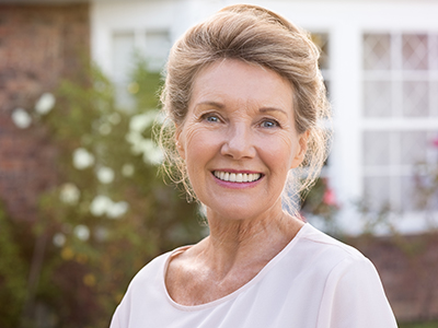 The image shows an elderly woman with short hair, wearing a white top and smiling at the camera. She is standing in front of a house with a brick exterior.