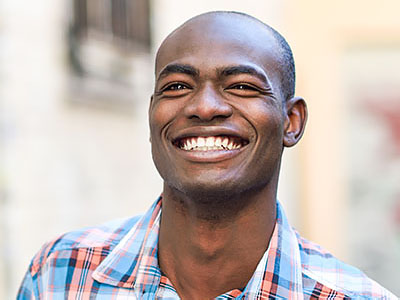 The image features a smiling man with short hair, holding his teeth in a playful manner. He is wearing a light blue shirt and has a beard. The background is out of focus but appears to be an urban setting with a building and a tree visible.