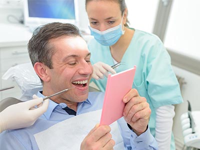 The image shows a man sitting in a dental chair, holding up a pink card with a smile on his face, surrounded by dental professionals who appear to be engaged in a discussion or presentation.