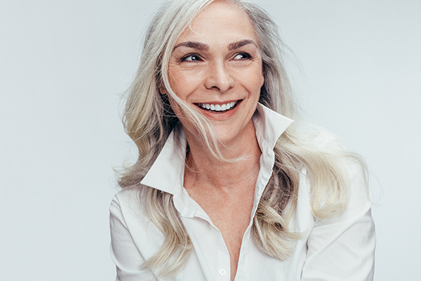 The image shows a woman with short blonde hair smiling at the camera. She has her hand on her chin and appears to be wearing a white top.