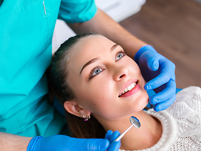 A dental hygienist is performing a teeth cleaning procedure, with the patient sitting in the chair and looking directly at the camera.