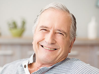 The image is a photograph of an elderly man with gray hair, wearing glasses and a blue shirt, smiling broadly at the camera while sitting in a relaxed posture.